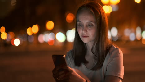 Nachts-Sitzt-Ein-Junges-Mädchen-Im-Gras-Einer-Großstadt,-Schaut-Auf-Den-Smartphone-Bildschirm,-Schreibt-Eine-Nachricht-Und-Pflegt-Ihr-Persönliches-Blog.